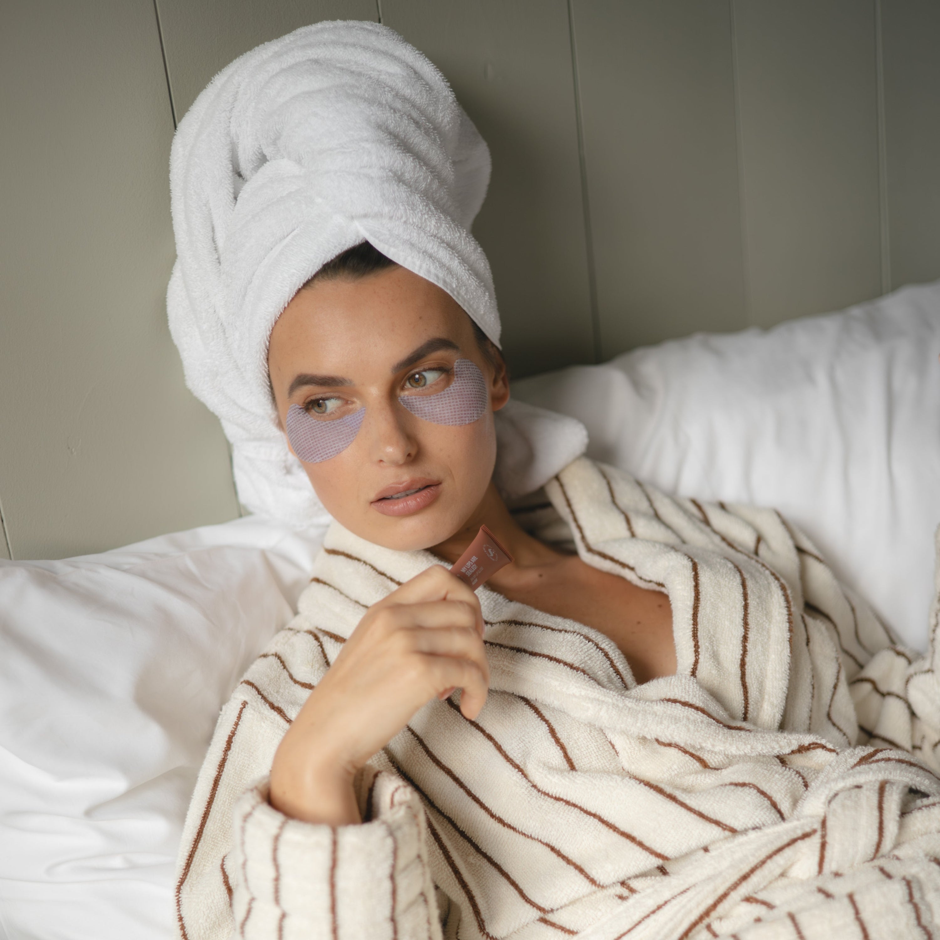Vrouw met badjas liggend op bed met oog patches onder haar ogen